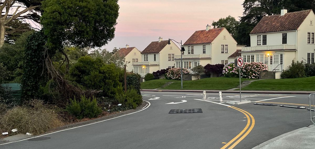 A quiet residential street at dusk: older homes with small front lawns under a fading pastel sky, one window already lit