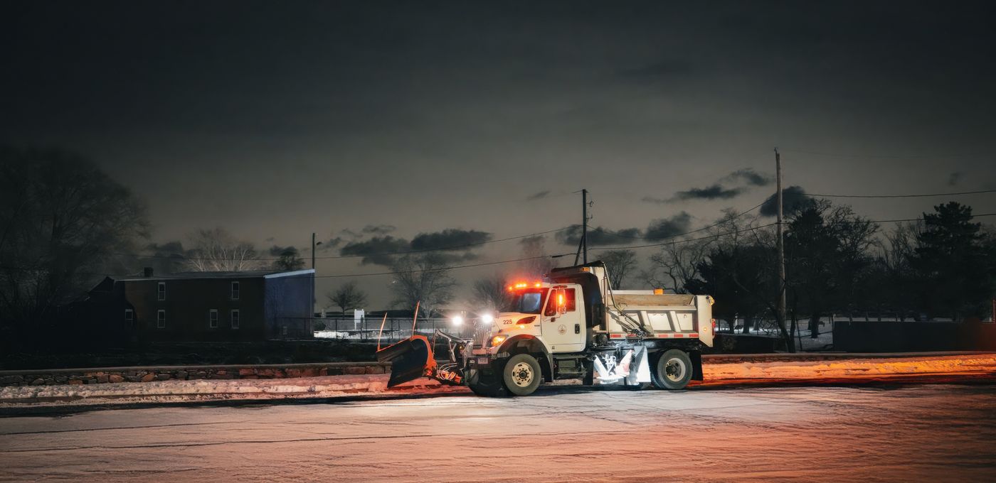 A plow truck working a snowy lot at night under warm streetlight
