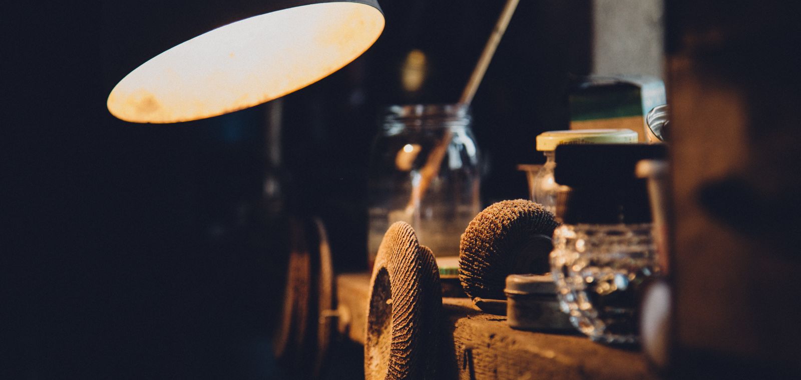 A workshop bench at night, lit by a single warm lamp over a wooden shelf of tools and jars